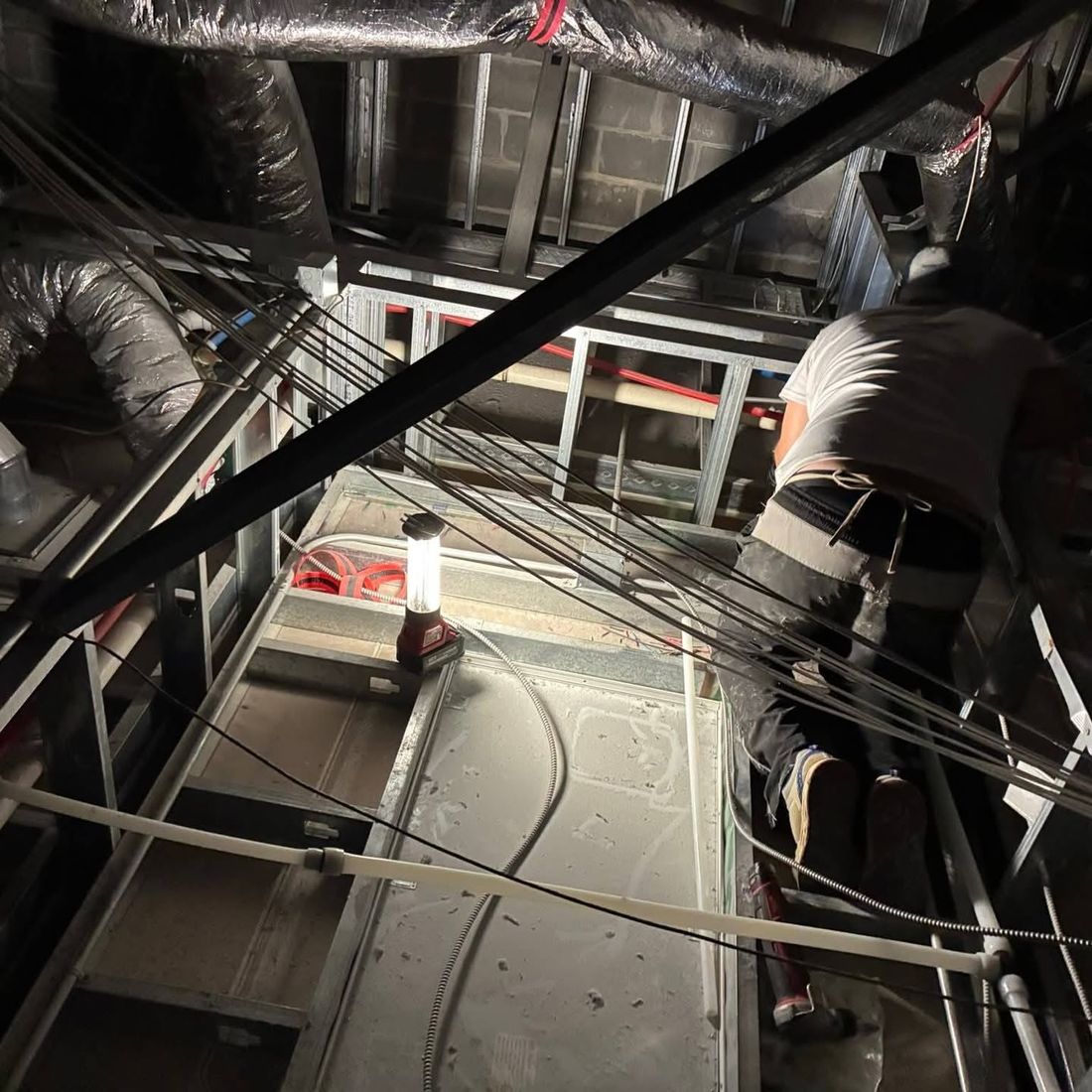 Overhead pipe and conduit being run in a commercial ceiling cavity, lit by a single work light.